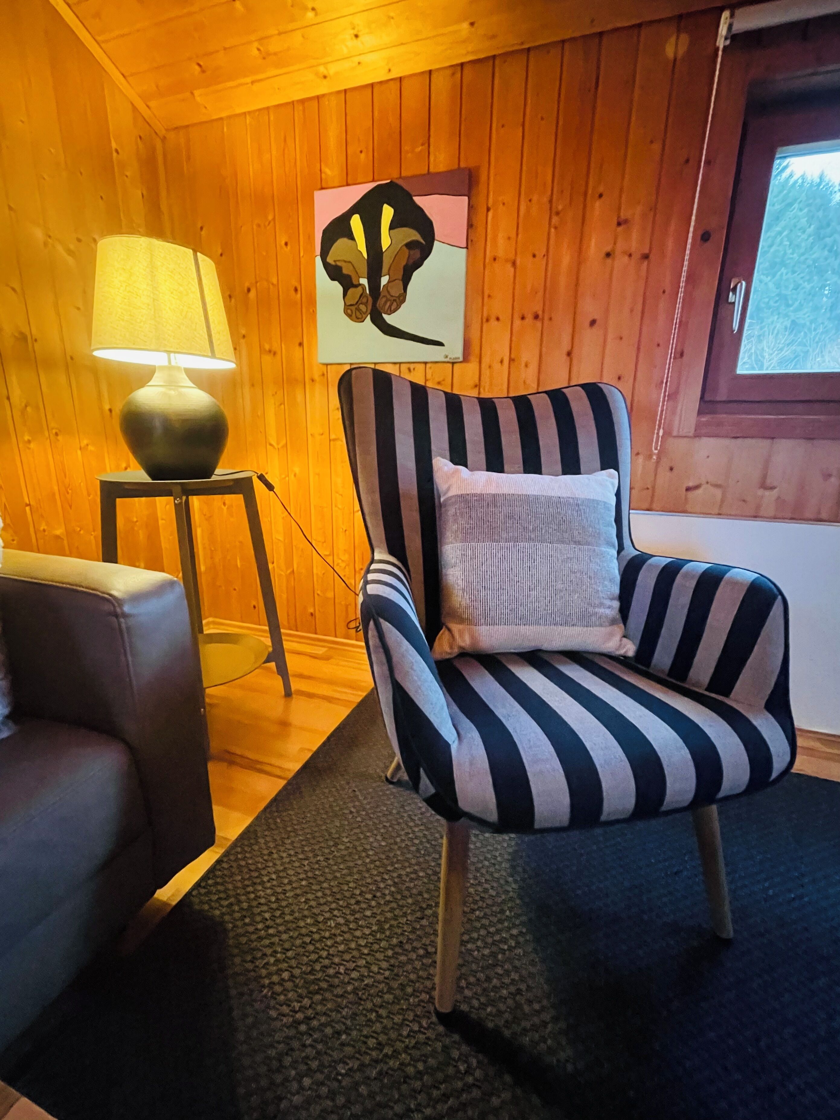 A cozy room with wooden walls, a striped armchair with a pillow, a small table with a lamp, and a painting on the wall.