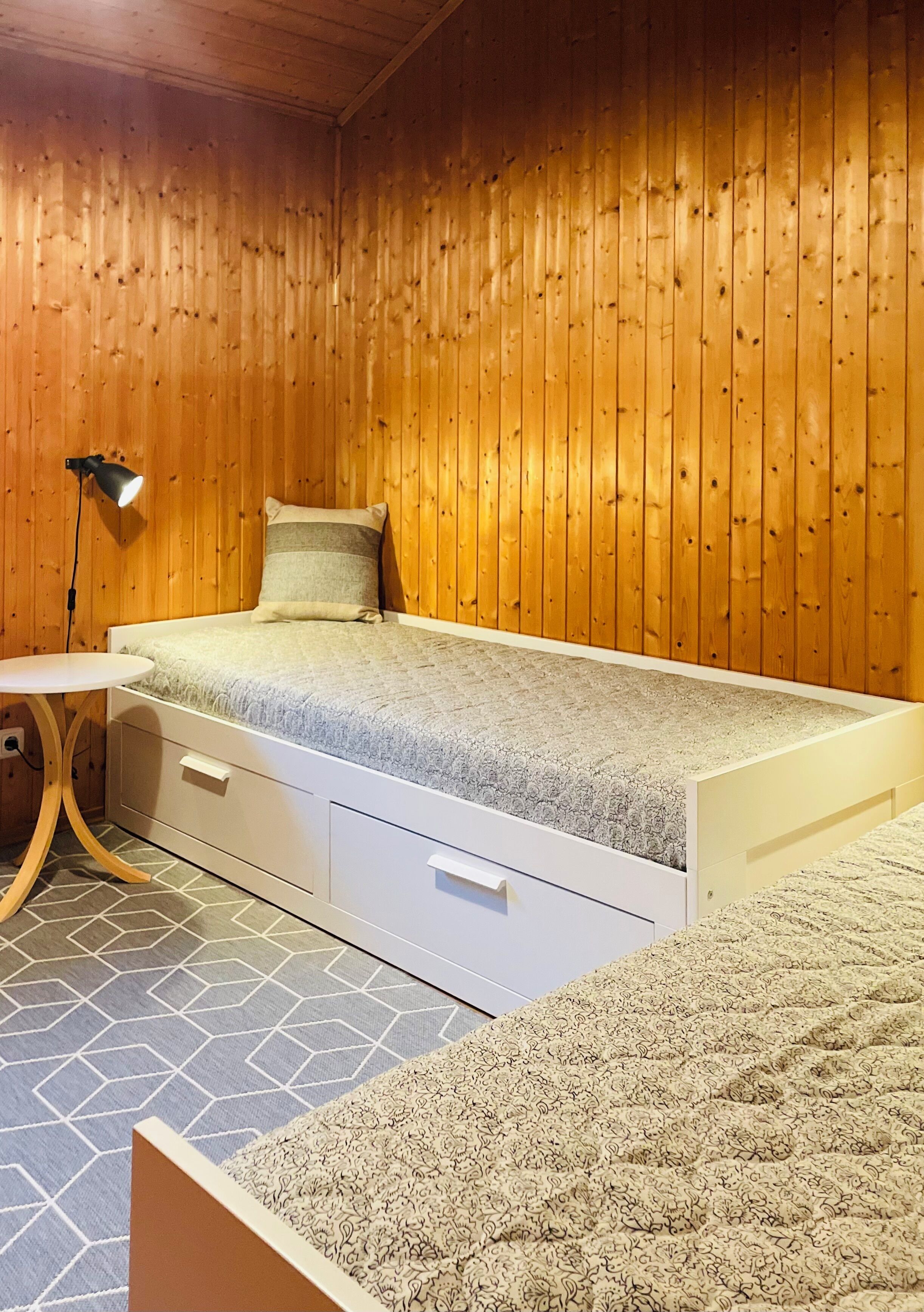 Una stanza con pareti in legno, un letto con struttura bianca, un piccolo comodino con una lampada e un tappeto decorato.