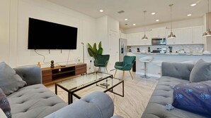 Ein modernes Wohnzimmer mit Fernseher, bequemen Sitzmöglichkeiten und einer Küche im Hintergrund.