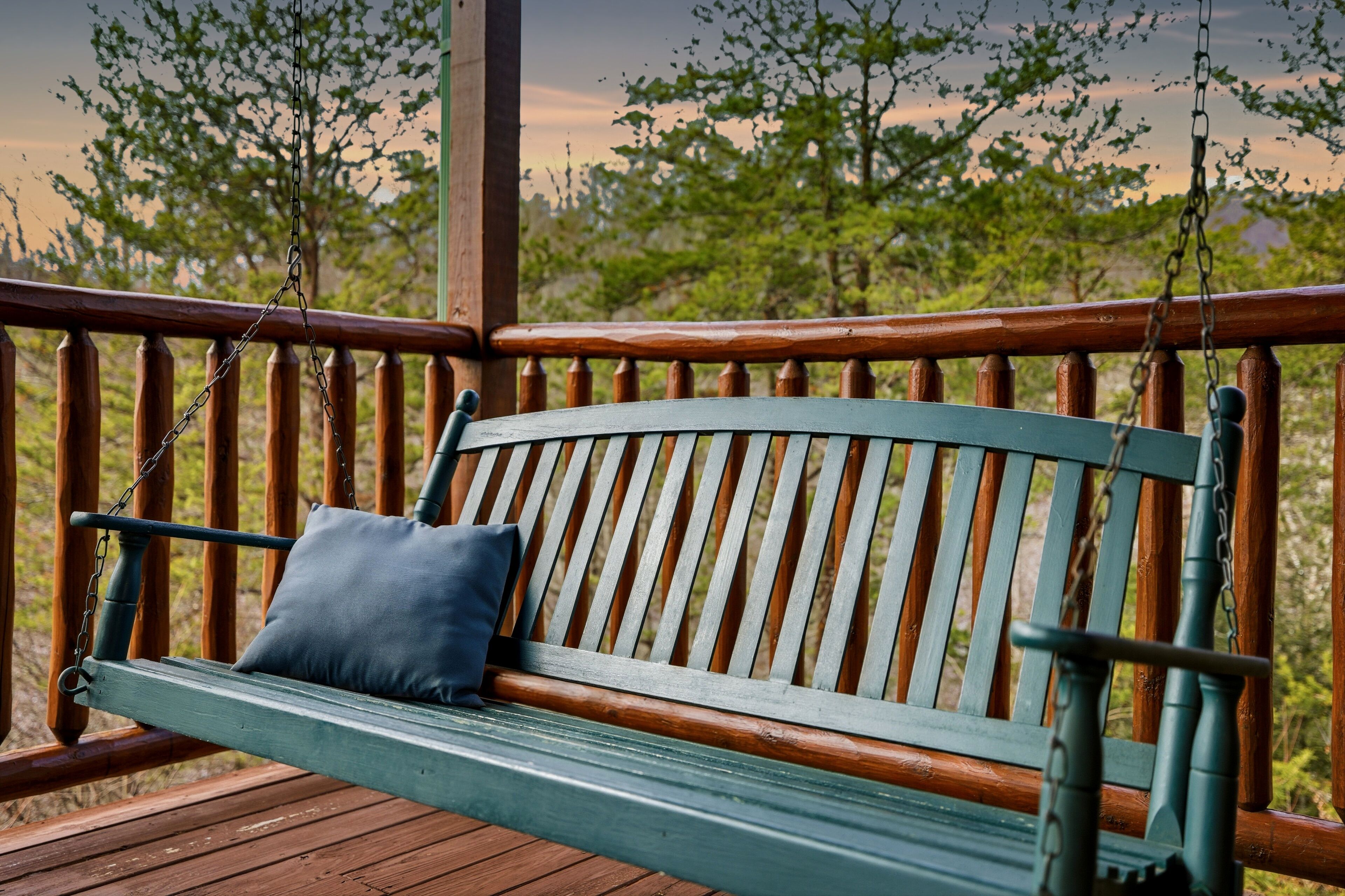A wooden swing bench with a cushion, attached to a wooden railing, overlooking a natural landscape.
