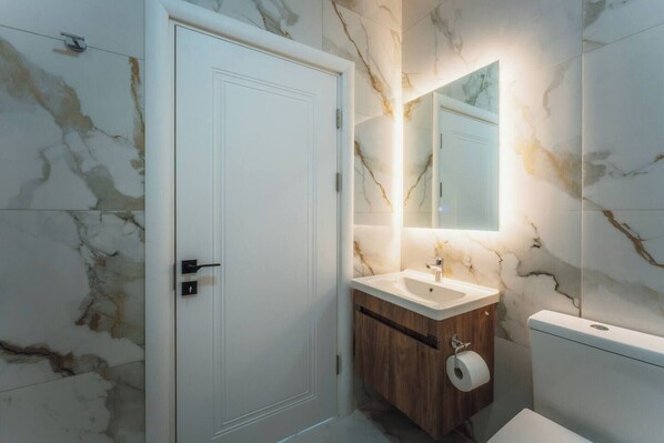 A bathroom with a marble wall, a wooden vanity, a white sink, a mirror, and a toilet. - Bright Apartment With Sea Valley Views BBQ (Nadur)