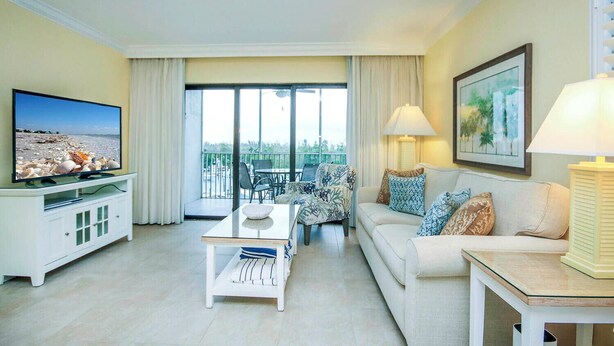 A spacious living room with a flat-screen TV, a sofa, a coffee table, and a balcony with a view of the beach.