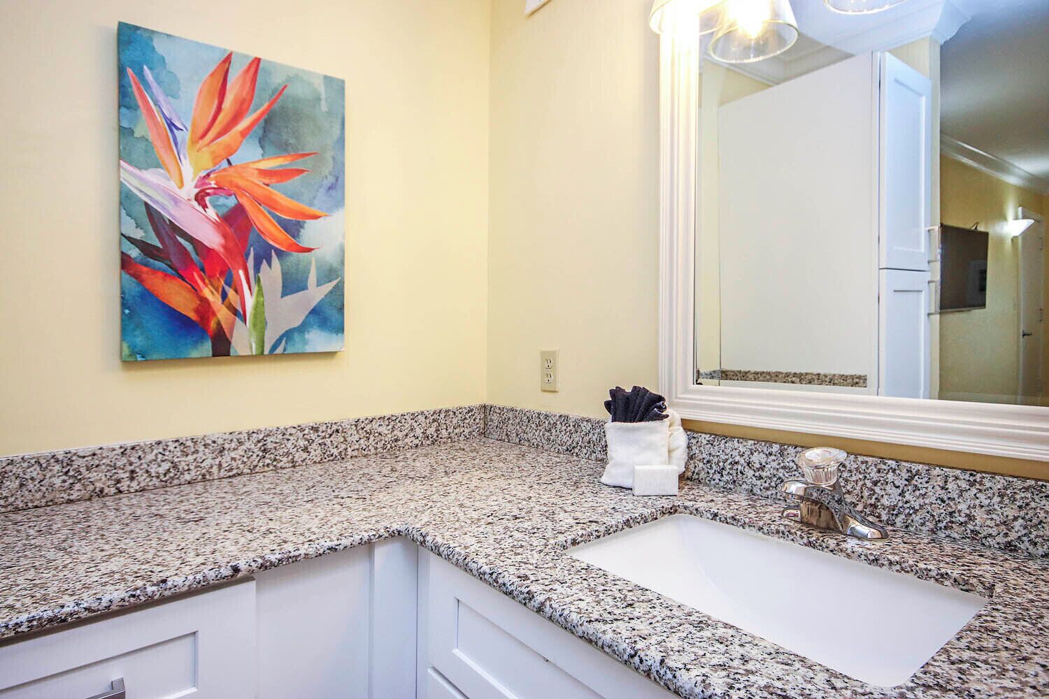 A bathroom with a granite countertop, a white sink, a large mirror, and a colorful painting on the wall.