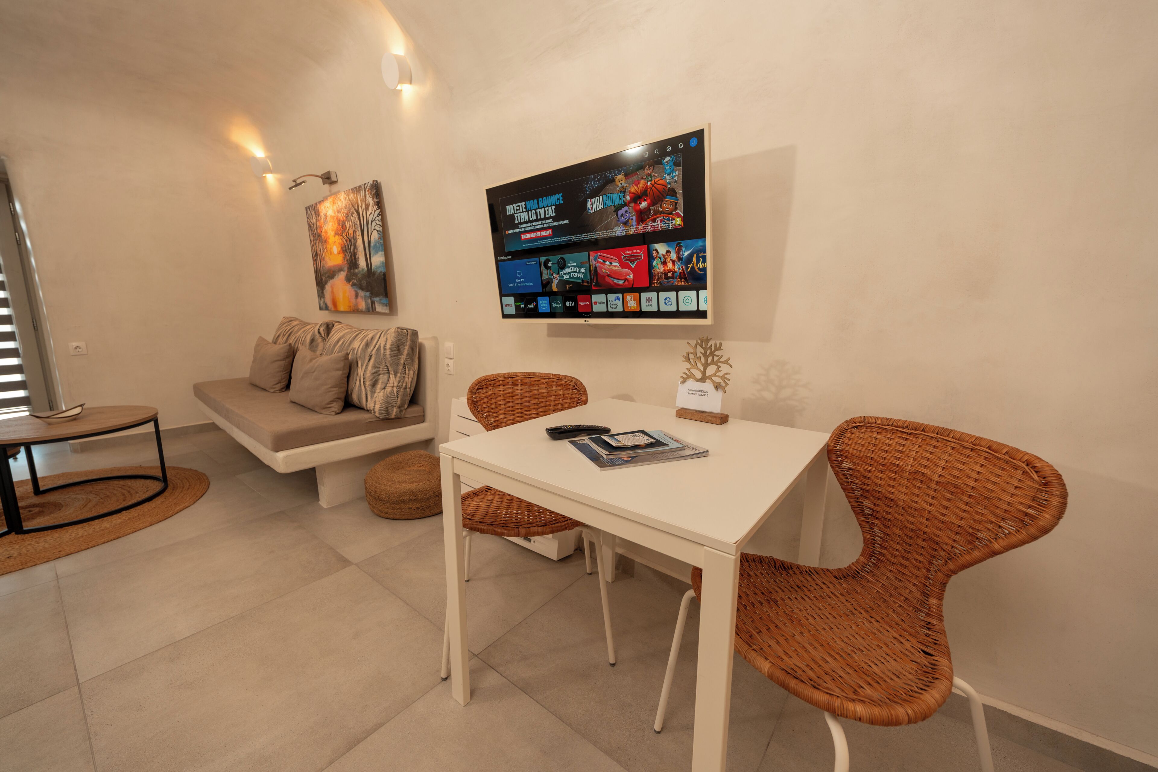 Una sala de estar moderna con un televisor de pantalla plana, un sofá, una mesa de comedor, sillas de mimbre y una pequeña planta decorativa.