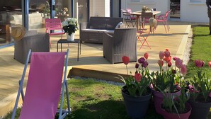 Et moderne hus med en patio med udendørs møbler, en have med tulipaner og et stort vindue med udsigt over haven.