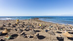 A beach with numerous thatched umbrellas and sun loungers, a lifeguard tower, and a rocky breakwater in the background. - Club Atlantis nice sea & pool view (Costa Adeje)