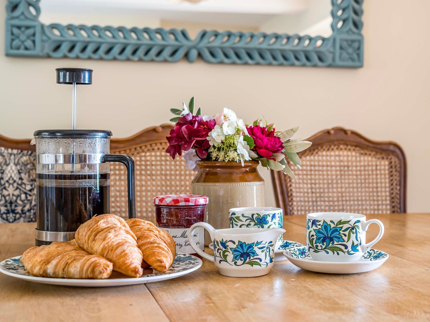 A breakfast setup with croissants, a coffee maker, a jar of jam, a vase with flowers, and a set of blue and white floral cups and saucers.