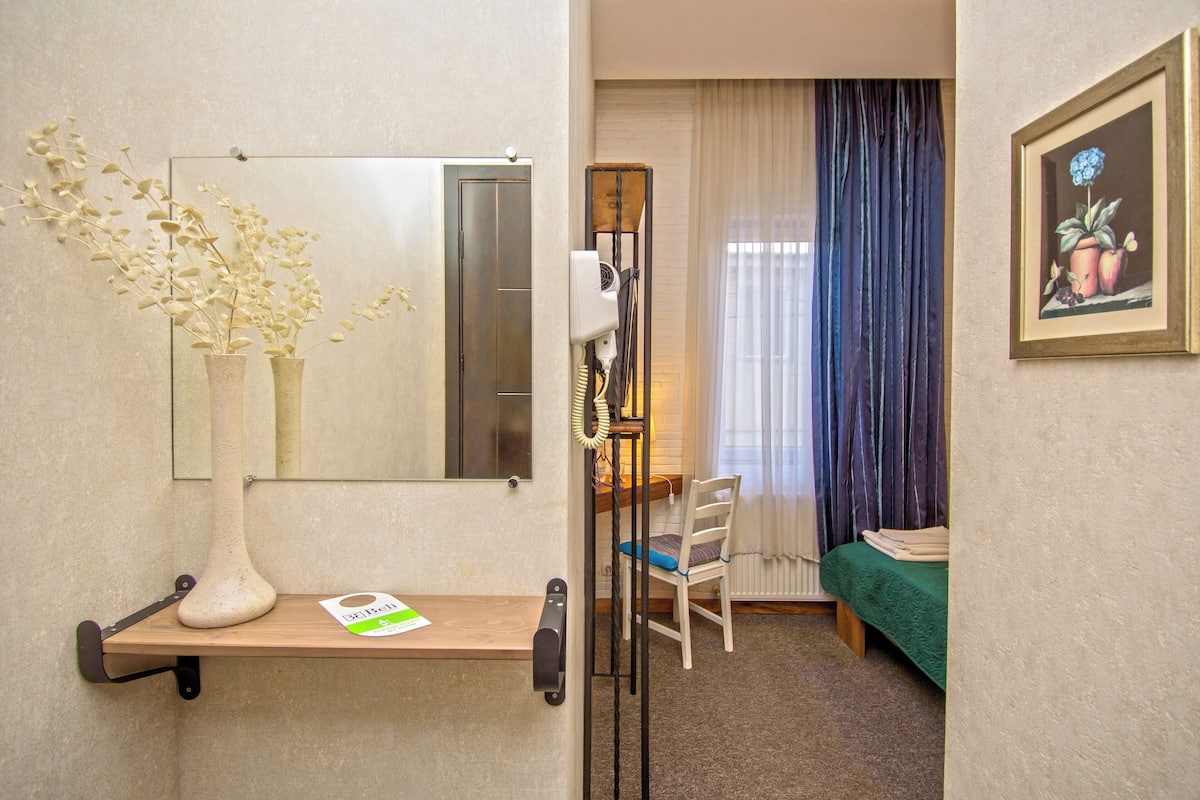 A hotel room with a wooden desk, a mirror, a vase with dried flowers, a chair, a bed with a green bedspread, and a framed picture on the wall.