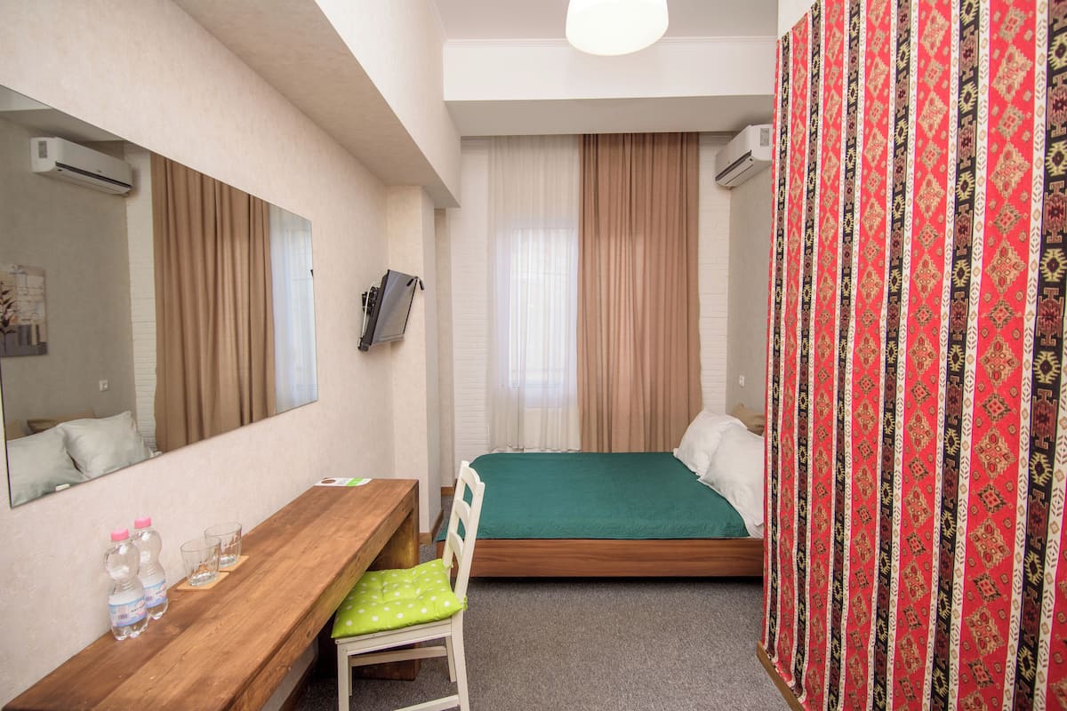 A hotel room with a wooden desk, a green bed, a chair, a mirror, and a curtain with a patterned design.