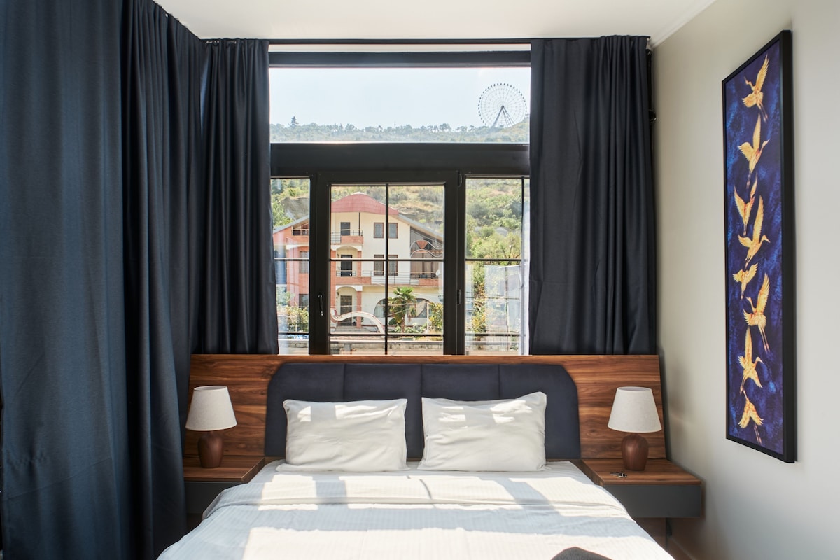 A hotel room with a large bed, two bedside tables with lamps, a window with curtains, and a wall-mounted artwork.
