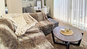 Ein Wohnzimmer mit einer Couch, einem Couchtisch und Blick auf eine Küche und einen Essbereich, der durch transparente Vorhänge sichtbar ist.