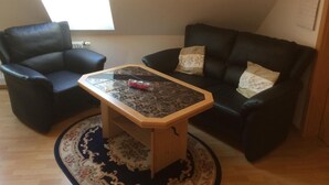 Een woonkamer met een salontafel met glazen blad, twee zwarte leren banken, een kamerkleed met patroon en een raam met natuurlijk licht.