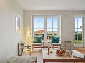Une pièce lumineuse et moderne, dotée d’une grande fenêtre, d’un coin salon confortable avec une table préparée pour le petit-déjeuner, et offrant une vue sur l’extérieur.