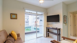 Ein Wohnzimmer mit einer Couch, einem Fernseher auf einem Schrank, einem Deckenventilator und einer Glastür zum Balkon.