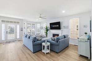 A living room with a sectional sofa, a fireplace, a television, and a ceiling fan. - Gold Coast 4BR Condo • Steps to Beach & Boardwalk (Ocean City)
