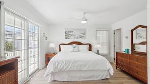 A bedroom with a large bed, a ceiling fan, a window with blinds, a dresser, and a wall-mounted mirror. - Gold Coast 4BR Condo • Steps to Beach & Boardwalk (Ocean City)