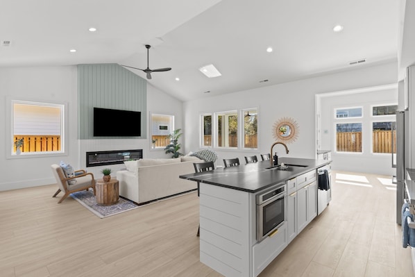 A modern kitchen with a central island, a dining area with a sofa and chairs, and a TV mounted on the wall. - Private Pool, Carolina Beach Rental, Dog Friendly, Luxury Finishes, Sleeps 9 (Carolina Beach)