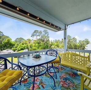 A covered outdoor seating area with a table and chairs, a floral-patterned floor, and a view of trees and buildings in the background. - The Wild Wahoo by Liquid Life (Orange Beach)