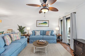 A living room with a blue sofa set, a coffee table, a ceiling fan, and a framed fish picture on the wall. - Cotton Bayou 7B by Liquid Life (Orange Beach)