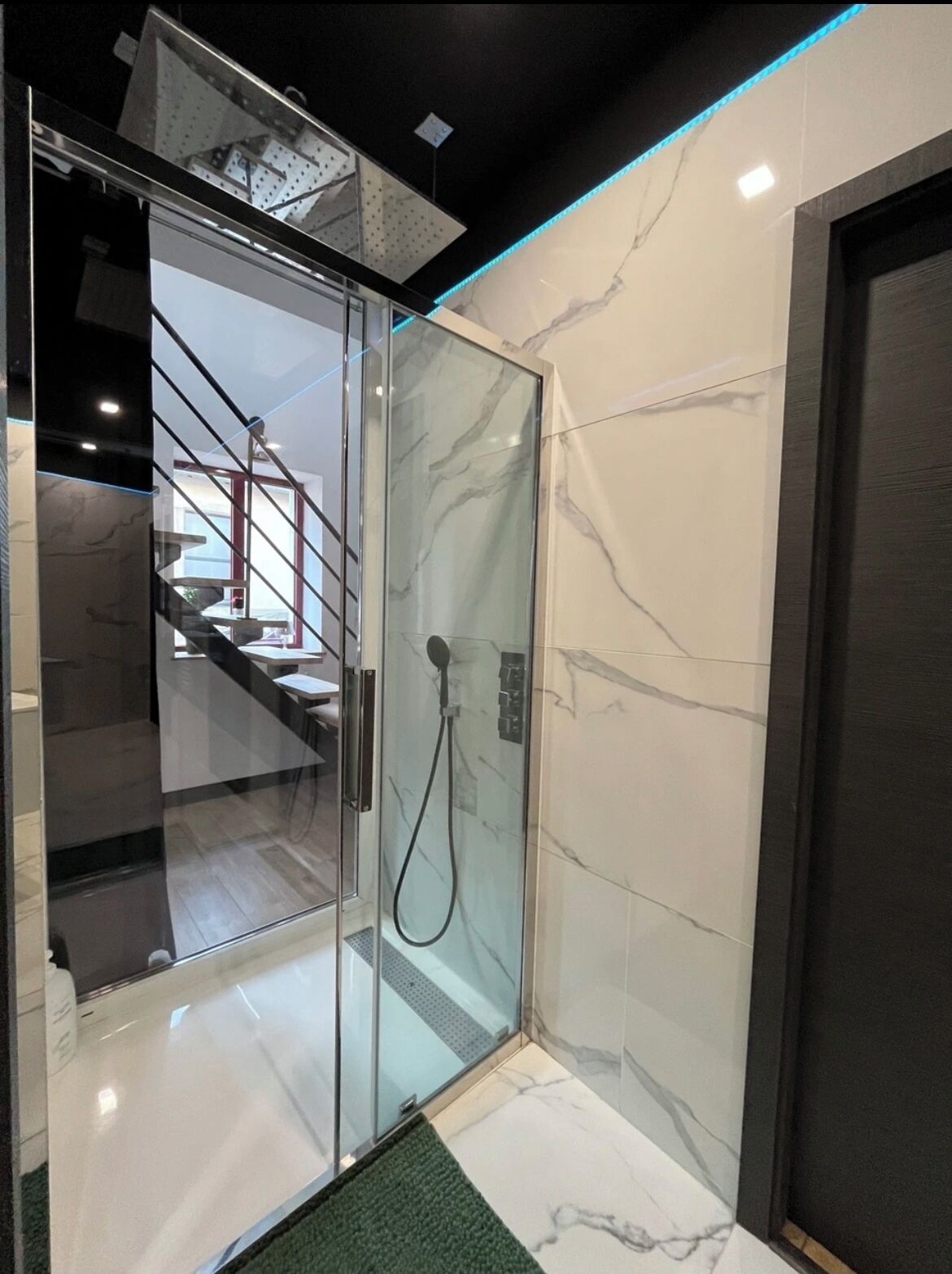 Ein modernes Badezimmer mit einer gläsernen Dusche, Marmorwänden und einer sichtbaren Treppe im Hintergrund.