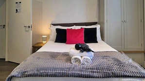 A neatly made bed with a grey blanket, a red pillow, and a black item on top. There is a small bedside table with a lamp.