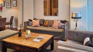 Un salon avec un canapé, une table basse et des lampes. Les murs sont ornés de tableaux encadrés et il y a une fenêtre avec des stores.