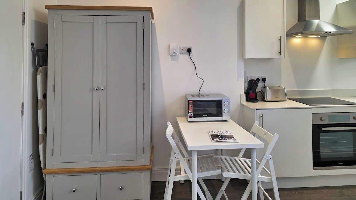 Eine kompakte Küche mit Mikrowelle, Toaster und Backofen. Ein kleiner Tisch mit einem Stuhl und eine Schrank mit Schubladen sind vorhanden.