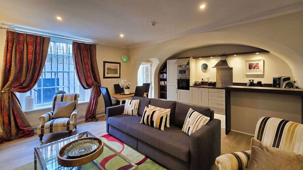A modern living room with a sofa, armchairs, and a coffee table. There is a kitchen area with a stove, microwave, and cabinets. The room has large windows with curtains and a view of the outside.