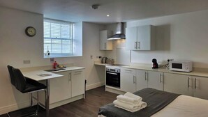 Cocina compacta con microondas, estufa y lavamanos. Zona de comedor pequeña con una silla y mesa. Se ve una cama con manta al frente.