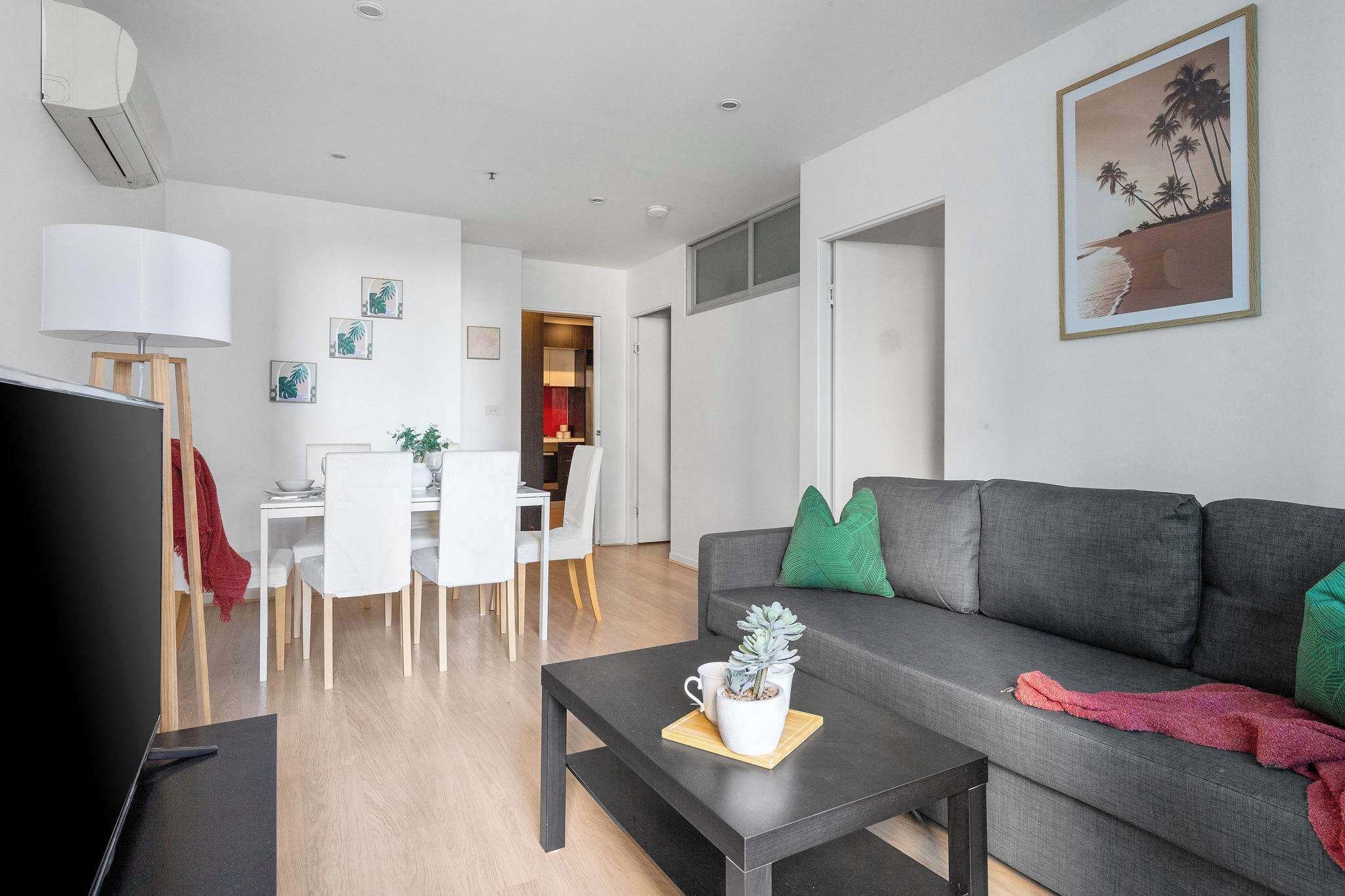 Un salon moderne avec un canapé gris, une table basse agrémentée d’une plante, un téléviseur et un coin repas avec des chaises blanches et un chemin de table rouge.
