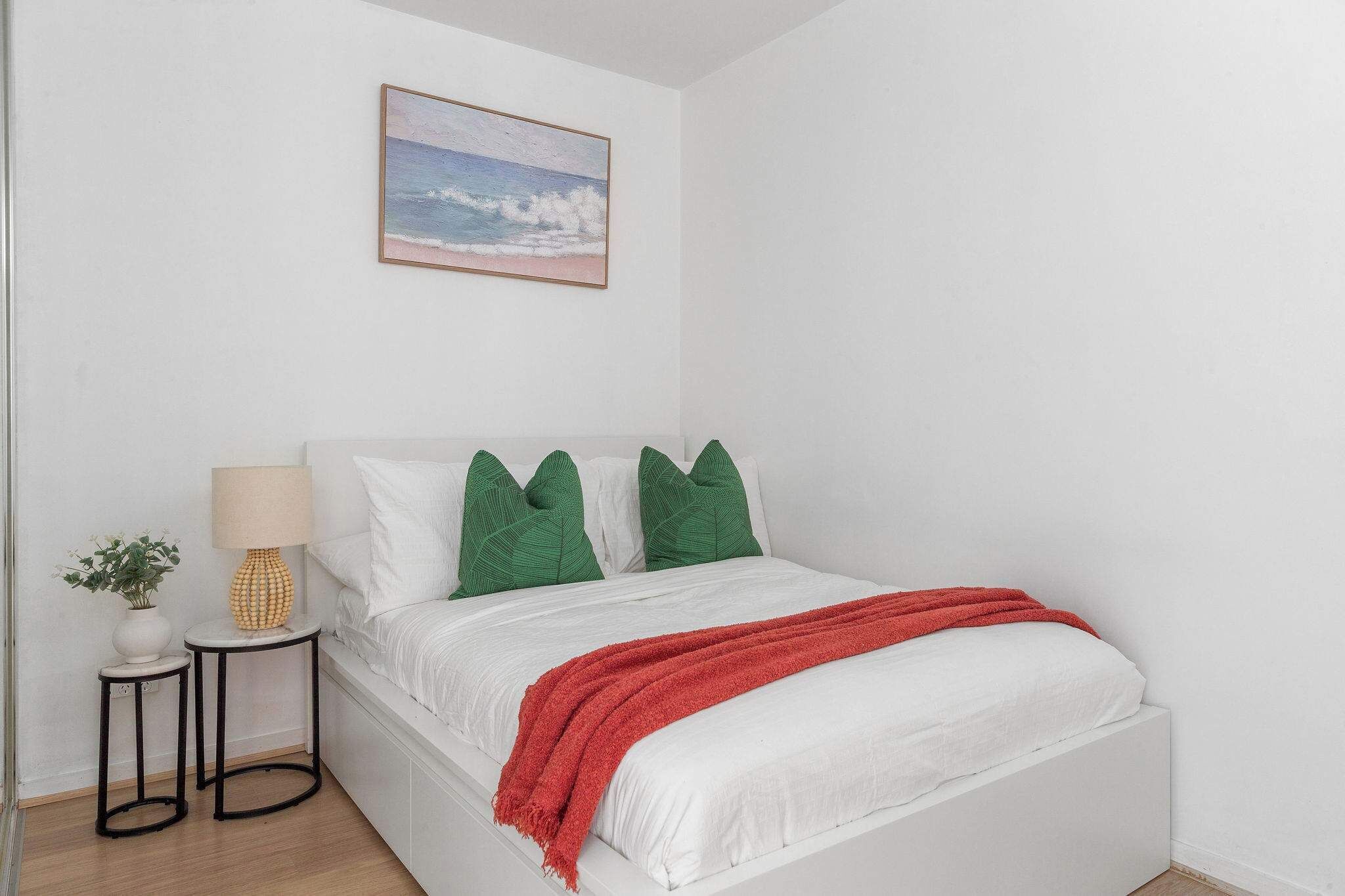 Cama bem arrumada com roupa de cama branca, almofadas verdes e manta vermelha. Uma pequena mesa com abajur e planta em vaso fica ao lado da cama.