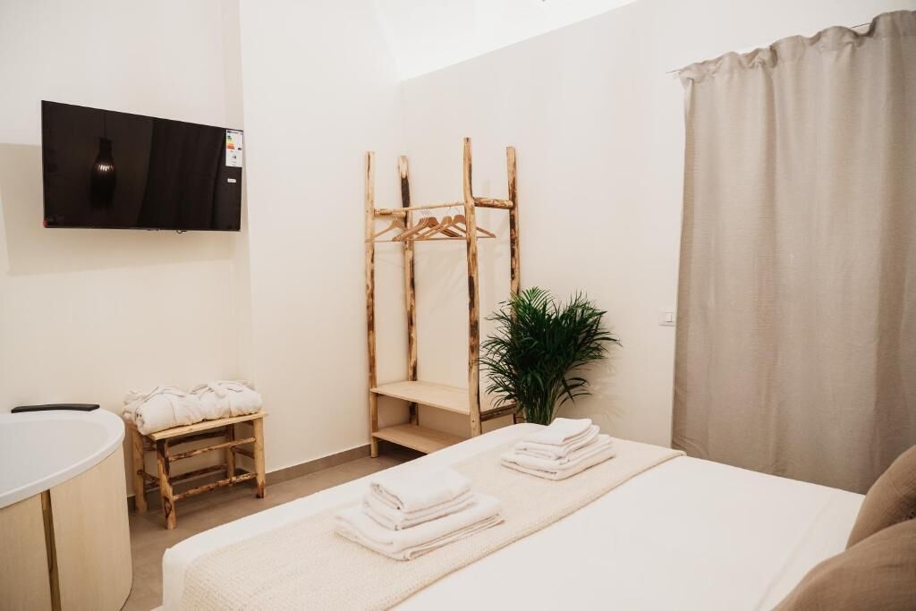 A modern hotel room with a bed, a wooden drying rack, a small stool, a plant, and a TV mounted on the wall.