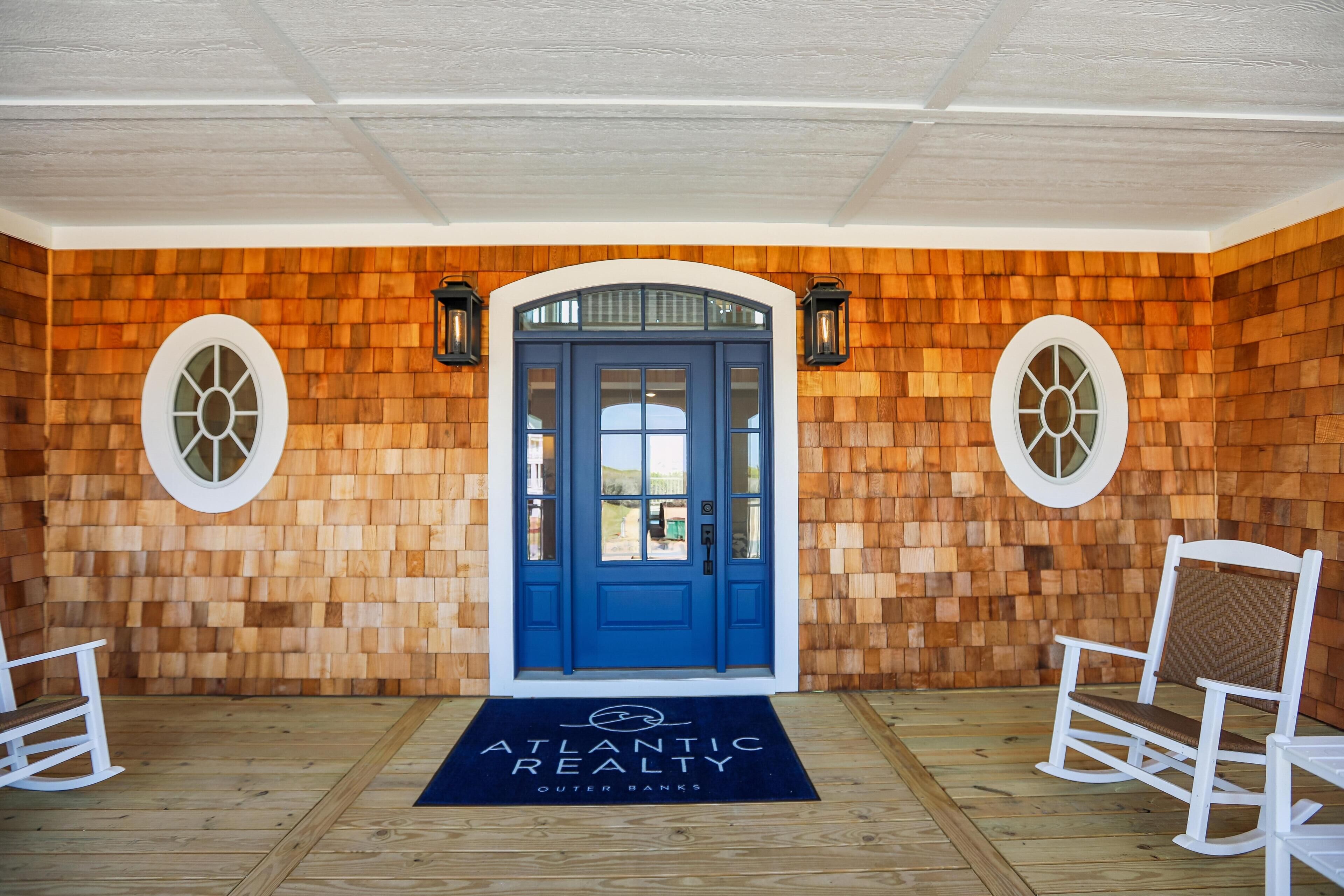 A wooden exterior wall with a blue door, two white rocking chairs, and a doormat with 'ATLANTIC REALTY' text.