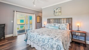 Une chambre à coucher avec un lit, un ventilateur de plafond, une table de chevet, une lampe et une vue sur un bâtiment à l’extérieur.