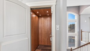 Un couloir avec un ascenseur en bois, une porte blanche et une fenêtre avec des stores.