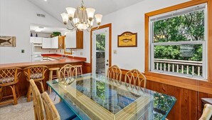 Un coin repas avec une table à plateau de verre, des chaises en osier, un bar en bois et une vue sur la verdure par une fenêtre.