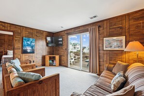 A cozy room with wooden paneling, a flat-screen TV, a sofa, a chair, and a table with a lamp. - Herons Return -- Flexible Arrival and Stay Dates Year Round (Kitty Hawk)