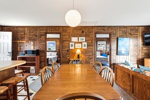 A dining area with a wooden table and chairs, a kitchen with a microwave, and a large window. - Herons Return -- Flexible Arrival and Stay Dates Year Round (Kitty Hawk)