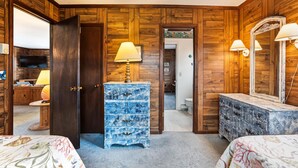 A room with wooden panel walls, a blue chest of drawers, a bed with floral bedding, and a mirror with wall sconces. - Herons Return -- Flexible Arrival and Stay Dates Year Round (Kitty Hawk)