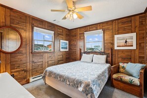 Ein Schlafzimmer mit Holzvertäfelung, einem Bett mit gemusterten Textilien, einem Sessel mit farbigem Kissen und zwei gerahmten Bildern an der Wand.