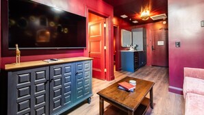 Una habitación con una televisión de pantalla grande, una mesa de centro de madera con libros, una puerta roja y una cocina al fondo.