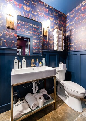 Ein Badezimmer mit blauer Wand, einem weißen Waschbecken, einer Toilette und einem Regal mit Handtüchern und Pflegeprodukten.