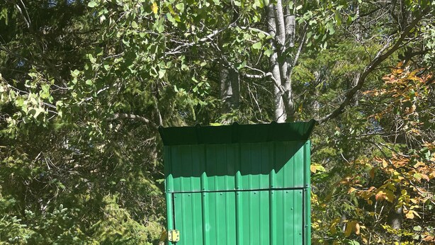 En grön metallbod med en dörr, placerad på en grund av betong, omgiven av träd och gräs.
