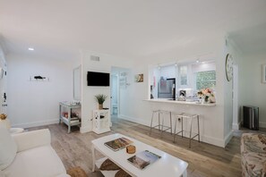 A modern living room with a white sofa, a coffee table with books, and a dining area with a kitchen in the background. - Bike to the Beach! Serene 1BR Sea Pines Condo / Pet Friendly! (Hilton Head Island)