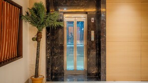 A lobby with a marble wall, a potted plant, and an elevator. - Hotel Prince Residency (Bhuj)