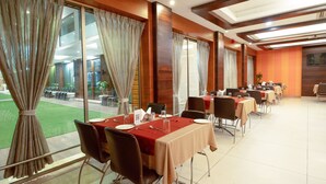 A hotel dining area with tables set for service, large windows with curtains, and a view of the outdoor area. - Hotel Prince Residency (Bhuj)