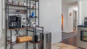 A kitchen with a metal shelf unit holding mugs, a coffee maker, and a toaster. There is a trash can and a wooden box on the floor.