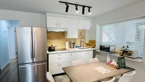 A modern kitchen with a stainless steel refrigerator, white cabinets, a wooden dining table with four chairs, and a window with blinds. - Metrotown sweet home 3-bedroom house in Burnaby perfect for your getaway (Burnaby)