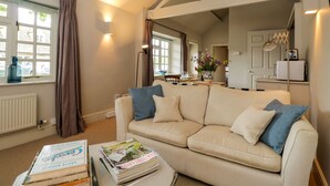 Ein Wohnzimmer mit einem beigefarbenen Sofa, einem Couchtisch mit Zeitschriften, einem Fenster mit Vorhängen und einer Küchenzeile im Hintergrund.