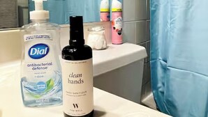 Un baño con un lavamanos blanco, una cortina de ducha azul y varios artículos de aseo, incluyendo una botella de jabón antibacterial Dial Defensa, una botella de desinfectante de manos The Well Limpieza de Manos y una botella de loción rosa.